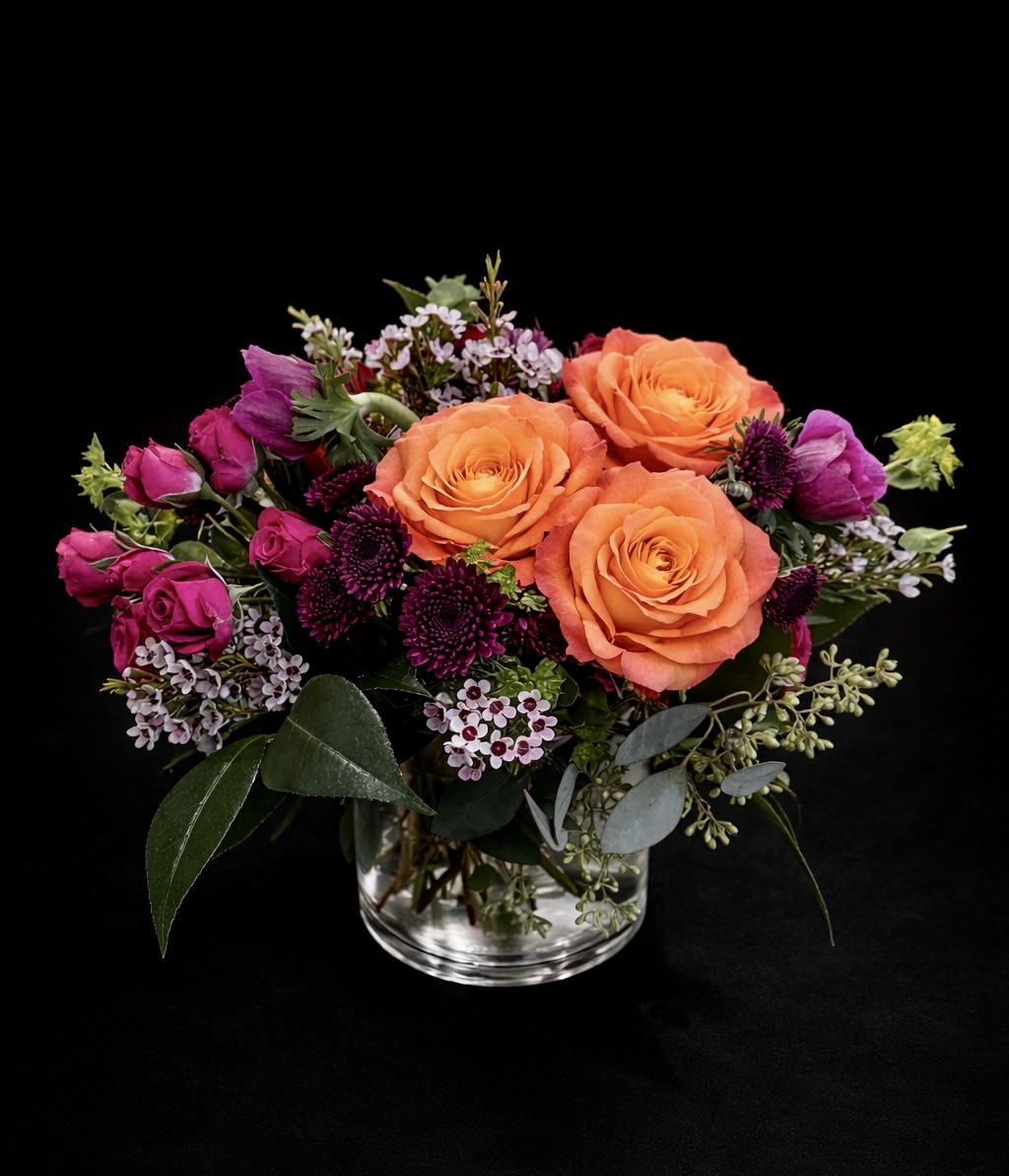 Luxury spring floral arrangement with orange roses, magenta spray roses, ranunculus, and waxflower in a glass vase on a black background by Vail Colorado florist A Secret Garden